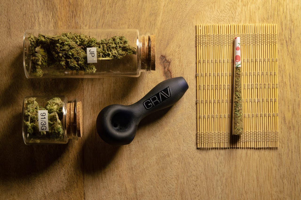 Glass jars with cannabis, a pipe, and a joint organized on a wooden tabletop.