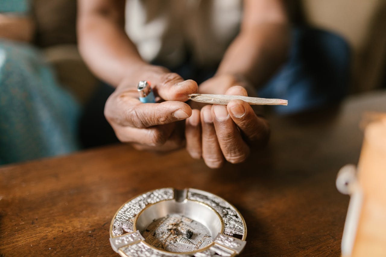 Close-up of hands holding a cannabis joint with an ashtray on a wooden table.