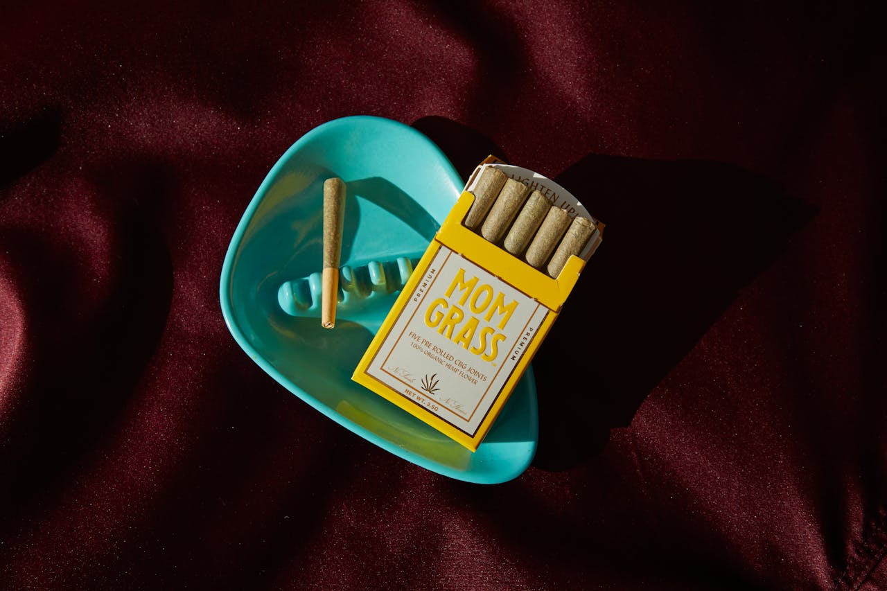 Flat lay of Mom Grass cannabis joints and blue ashtray on red fabric background, creative product display.